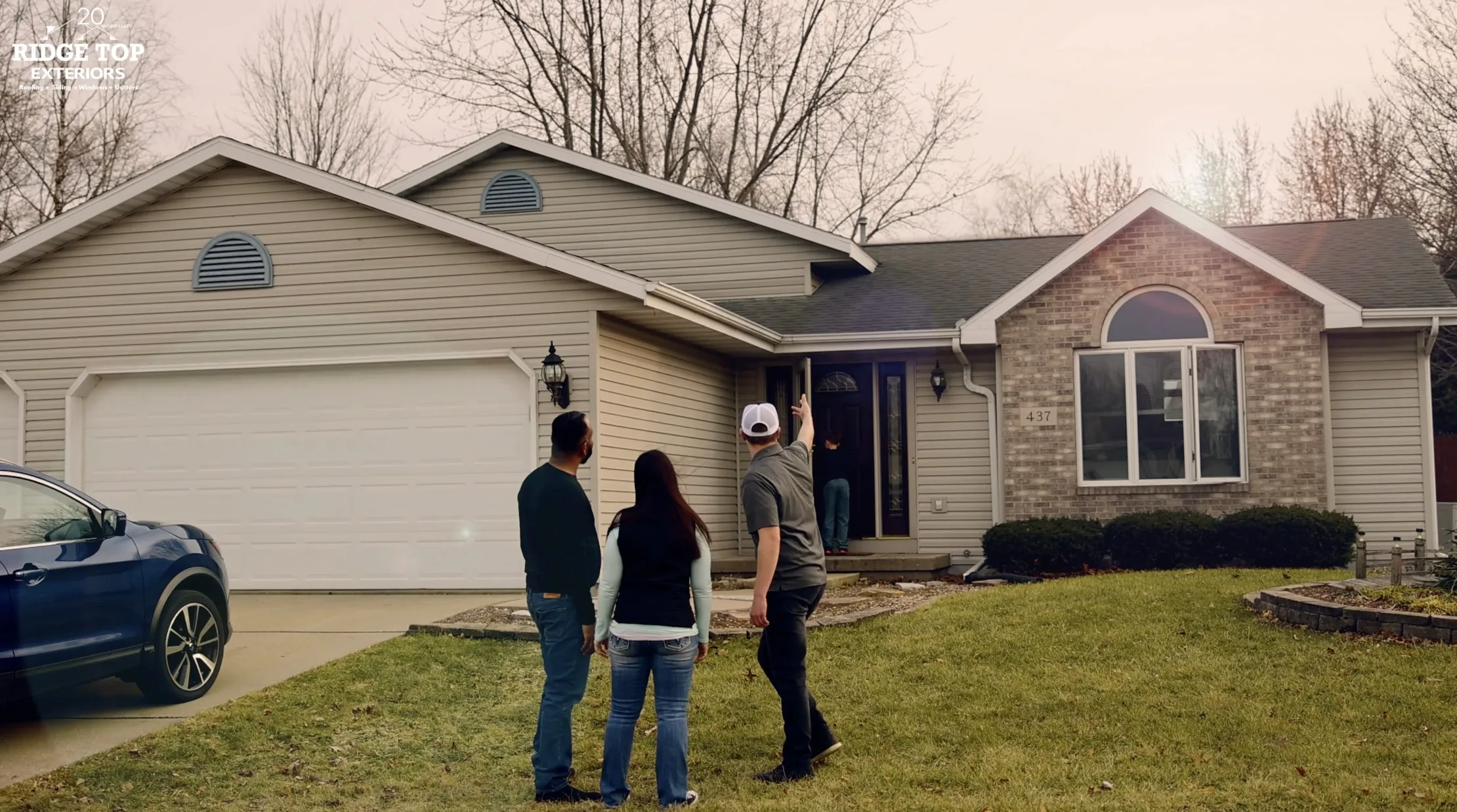 Three people standing on a lawn looking at a beige house with a blue car parked in the driveway and a child entering the front door.